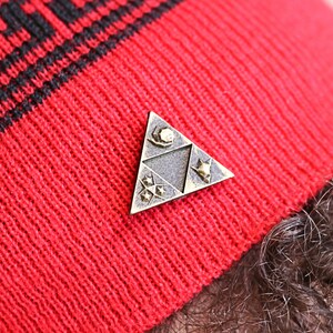 May include: A red knit beanie with a black and white striped pattern. A gold-colored triangular pin with a design of a stylized triangle with a crescent moon, a star, and a triforce symbol is attached to the beanie.