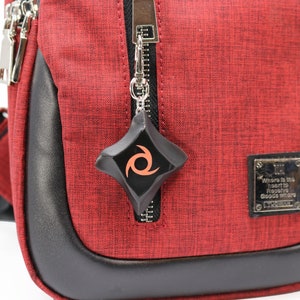 May include: A red crossbody bag with a black zipper and a black leather trim. A black and orange ninja star keychain is attached to the zipper. The bag has a small silver patch with the text "Where is the heart to fight? Good where good is."