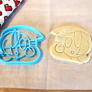 May include: A blue plastic cookie cutter shaped like a cartoon character with a hat, next to a light brown cookie that has been cut with the cutter.
