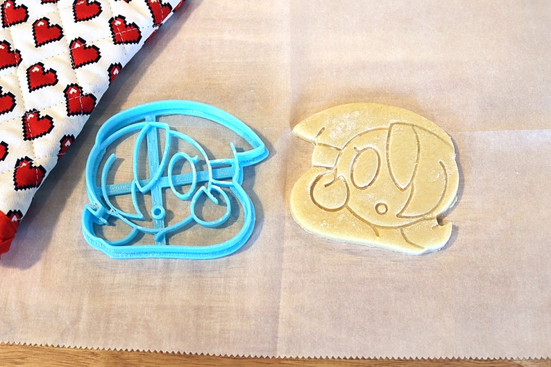 May include: A blue plastic cookie cutter shaped like a cartoon character with a hat, next to a light brown cookie that has been cut with the cutter.
