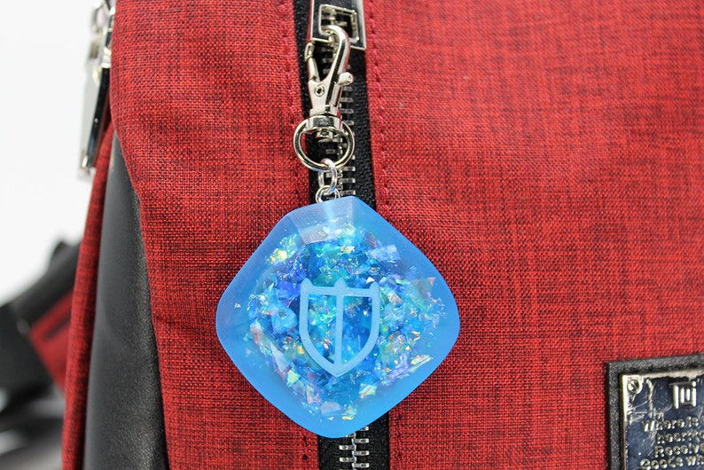 May include: A blue resin charm with a silver shield design and iridescent glitter inside. The charm is attached to a silver zipper pull.