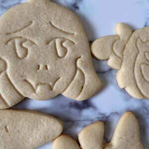 May include: Five sugar cookies decorated with various designs. The cookies are light brown and have a slightly rough texture. One cookie features a cartoon face with large eyes and a sad expression. Another cookie has a design of a creature with large eyes and a mouth.