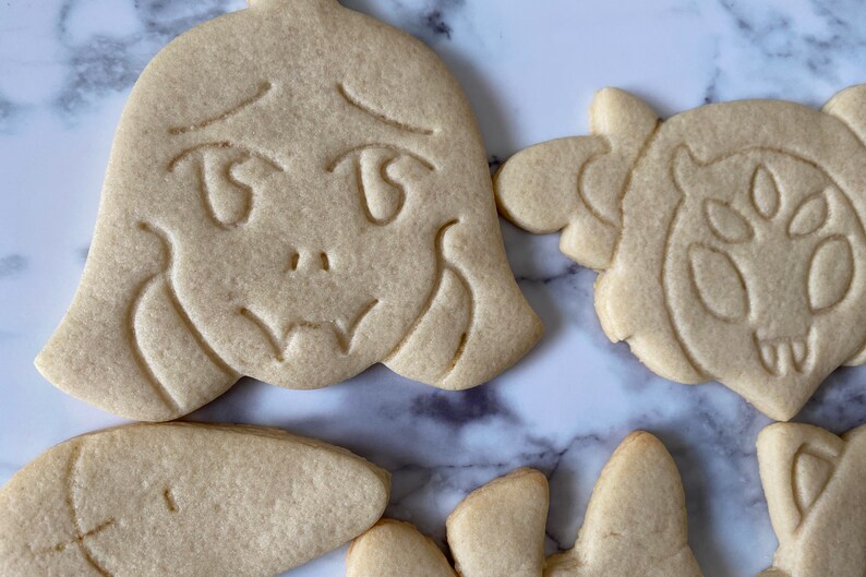 May include: Five sugar cookies decorated with various designs. The cookies are light brown and have a slightly rough texture. One cookie features a cartoon face with large eyes and a sad expression. Another cookie has a design of a creature with large eyes and a mouth.