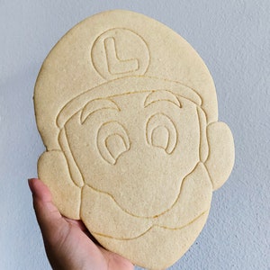 May include: A large, light brown cookie shaped like the face of a cartoon character, with a hat and the letter "L" on it. The cookie has detailed facial features and is held in a hand.