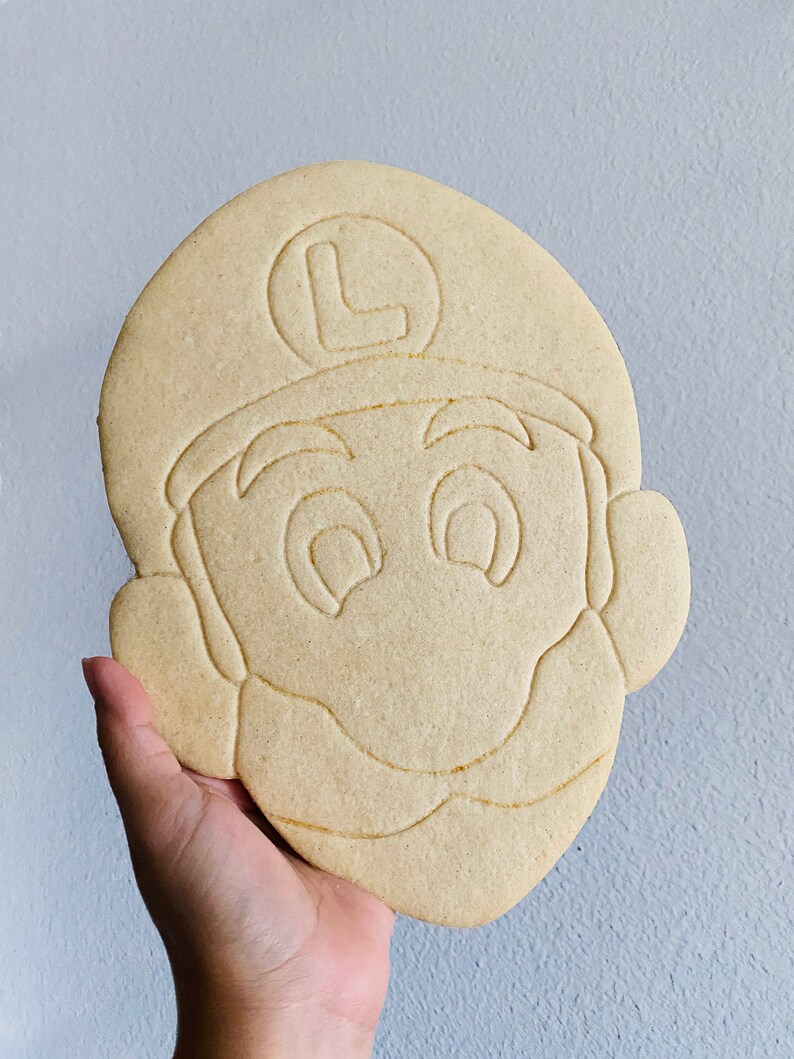 May include: A large, light brown cookie shaped like the face of a cartoon character, with a hat and the letter "L" on it. The cookie has detailed facial features and is held in a hand.