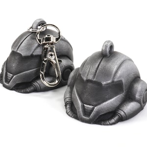 Puede incluir: Dos llaveros en forma de casco de color plateado con un anillo y un cierre de metal plateado. Los llaveros están detallados con superficies texturizadas y tienen un diseño futurista.