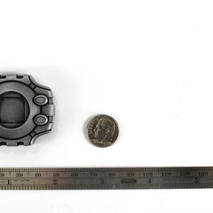 Puede incluir: Una r&eacute;plica de metal plateado de un dispositivo Digimon, una consola de videojuegos port&aacute;til popular, con una pantalla cuadrada y un bot&oacute;n circular en el lateral. El dispositivo tiene un di&aacute;metro de aproximadamente 5 cm y est&aacute; sobre una superficie blanca.