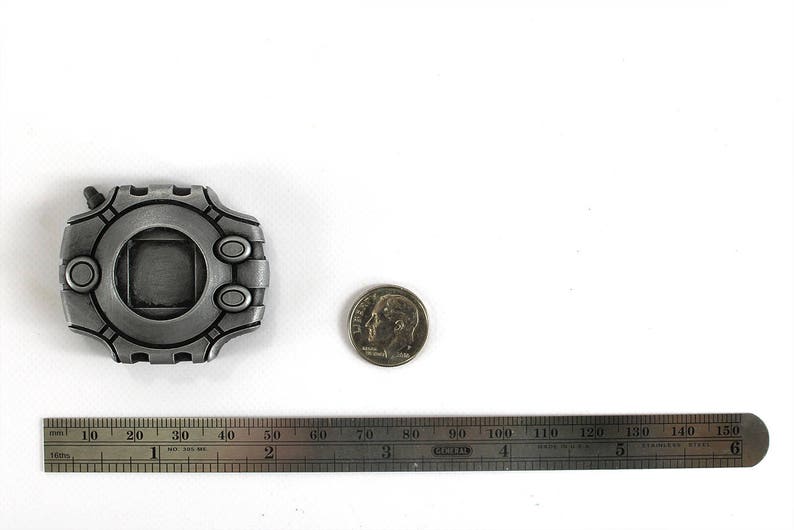 Puede incluir: Una r&eacute;plica de metal plateado de un dispositivo Digimon, una consola de videojuegos port&aacute;til popular, con una pantalla cuadrada y un bot&oacute;n circular en el lateral. El dispositivo tiene un di&aacute;metro de aproximadamente 5 cm y est&aacute; sobre una superficie blanca.