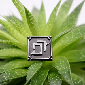 May include: A silver-colored square pin with a stylized Japanese character in the center. The pin is sitting on a green succulent plant.