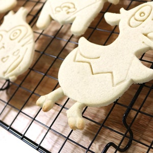 May include: A close-up of a cooling rack with several sugar cookies. The cookies are shaped like cartoon characters and are un-decorated.