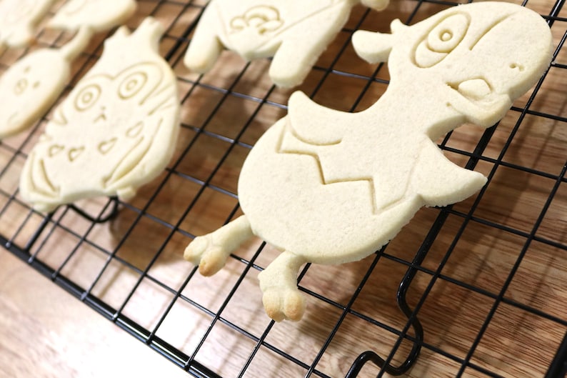 May include: A close-up of a cooling rack with several sugar cookies. The cookies are shaped like cartoon characters and are un-decorated.
