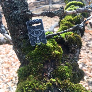 May include: A silver metal charm featuring a stylised eye design and geometric patterns. The charm is hanging from a tree branch covered in moss.