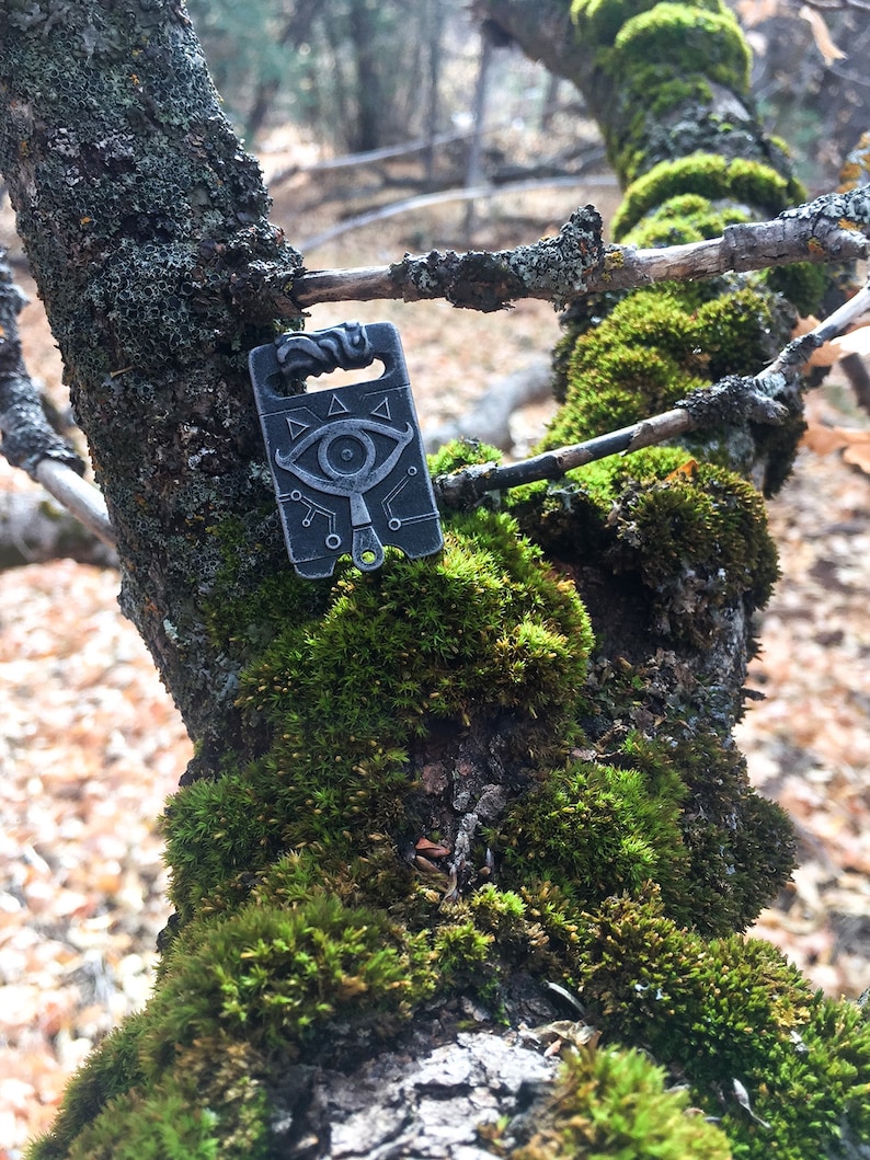 May include: A silver metal charm featuring a stylised eye design and geometric patterns. The charm is hanging from a tree branch covered in moss.