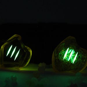 May include: Two glowing, amber-colored, organic-shaped objects with three bright green, vertical slashes. The objects are set against a dark background, with a subtle green glow emanating from the base.