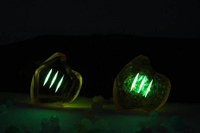 May include: Two glowing, amber-colored, organic-shaped objects with three bright green, vertical slashes. The objects are set against a dark background, with a subtle green glow emanating from the base.