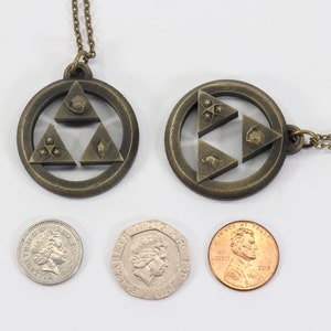 Puede incluir: Dos collares de color dorado con colgantes que representan el s&iacute;mbolo Triforce de la serie de videojuegos The Legend of Zelda. Los colgantes est&aacute;n hechos de metal y tienen un dise&ntilde;o detallado. Los collares se muestran junto a tres monedas para la escala.