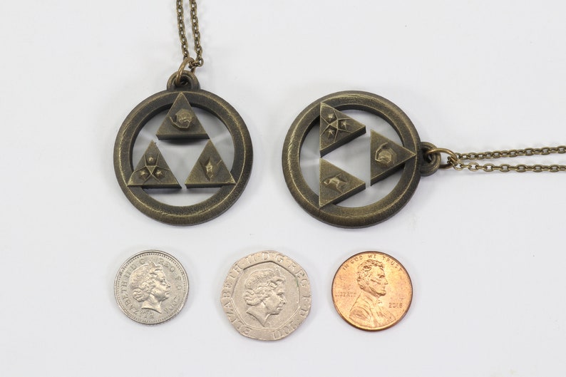 Puede incluir: Dos collares de color dorado con colgantes que representan el s&iacute;mbolo Triforce de la serie de videojuegos The Legend of Zelda. Los colgantes est&aacute;n hechos de metal y tienen un dise&ntilde;o detallado. Los collares se muestran junto a tres monedas para la escala.