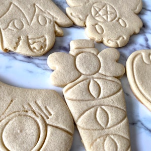 Pode incluir: Cinco biscoitos de a&ccedil;&uacute;car decorados com v&aacute;rios desenhos. Os biscoitos est&atilde;o sobre uma superf&iacute;cie de m&aacute;rmore branco. Os desenhos incluem um rosto, uma garrafa, um cora&ccedil;&atilde;o e outros dois desenhos.