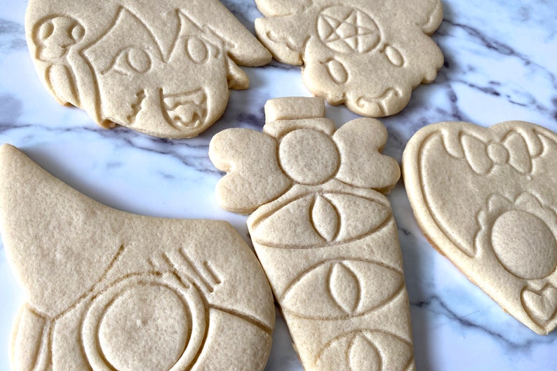 Pode incluir: Cinco biscoitos de a&ccedil;&uacute;car decorados com v&aacute;rios desenhos. Os biscoitos est&atilde;o sobre uma superf&iacute;cie de m&aacute;rmore branco. Os desenhos incluem um rosto, uma garrafa, um cora&ccedil;&atilde;o e outros dois desenhos.