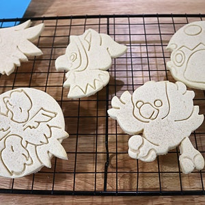 May include: Five unbaked sugar cookies on a black wire cooling rack. The cookies are shaped like different cartoon characters, including a bird, a dinosaur, and a mushroom.