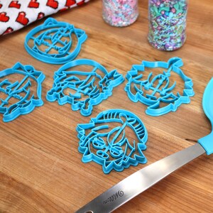 Pu&ograve; includere: Cinque tagliabiscotti in plastica blu a forma di personaggi dei cartoni animati su una superficie di legno. C'&egrave; anche una spatola blu con manico argentato sulla superficie.