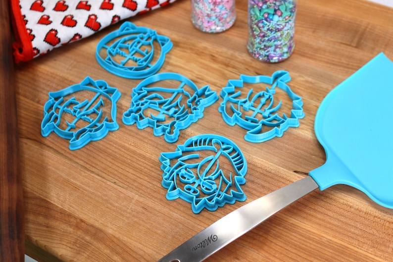 Pu&ograve; includere: Cinque tagliabiscotti in plastica blu a forma di personaggi dei cartoni animati su una superficie di legno. C'&egrave; anche una spatola blu con manico argentato sulla superficie.