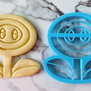 May include: A blue plastic cookie cutter shaped like a flower with a face. The flower has a round center with two eyes and a mouth, and two leaves at the bottom.