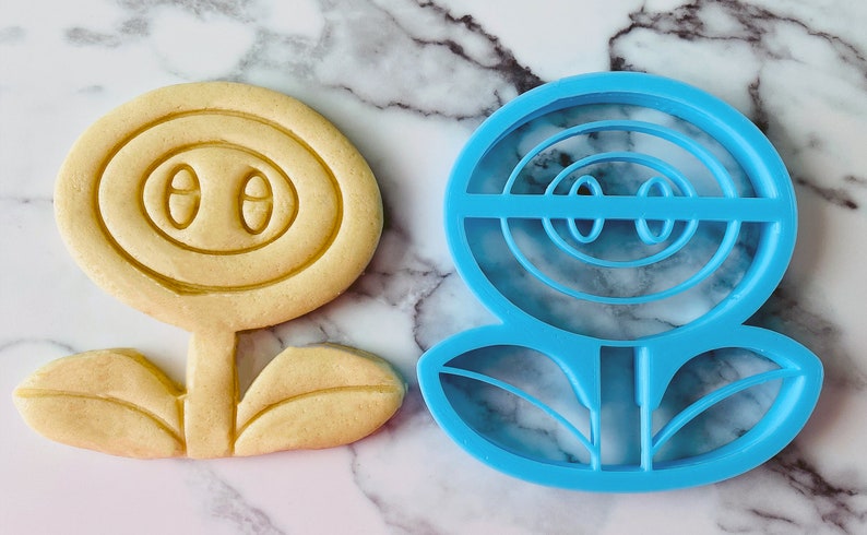 May include: A blue plastic cookie cutter shaped like a flower with a face. The flower has a round center with two eyes and a mouth, and two leaves at the bottom.