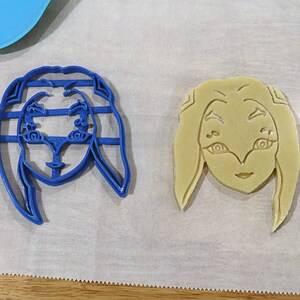May include: Two cookie cutters, one blue and one beige, shaped like a female face with long hair. The blue cutter is on a white surface, while the beige cutter is on a white surface with a brown wooden surface in the background.