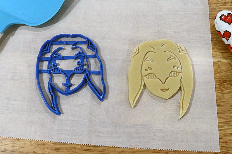 May include: Two cookie cutters, one blue and one beige, shaped like a female face with long hair. The blue cutter is on a white surface, while the beige cutter is on a white surface with a brown wooden surface in the background.