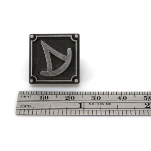 May include: A silver-colored metal pin with a square design. The pin features a stylized letter "D" in a silver color against a black background. The pin is approximately 2 cm in size.