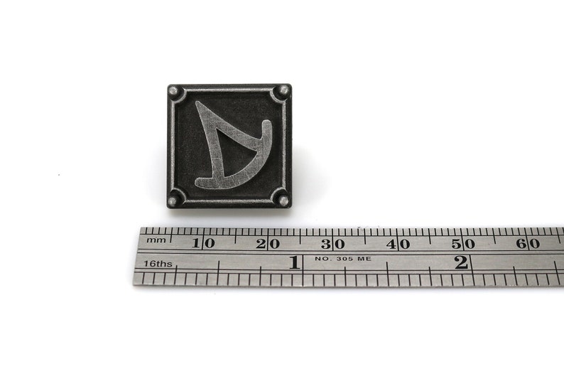 May include: A silver-colored metal pin with a square design. The pin features a stylized letter "D" in a silver color against a black background. The pin is approximately 2 cm in size.
