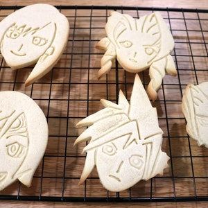 May include: Four decorated sugar cookies on a black wire cooling rack. The cookies are shaped like faces with different hairstyles and expressions.