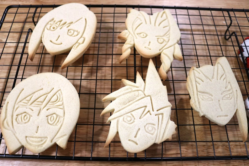 May include: Four decorated sugar cookies on a black wire cooling rack. The cookies are shaped like faces with different hairstyles and expressions.