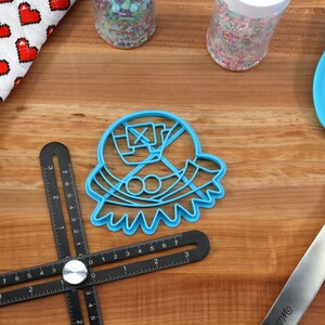 May include: A blue plastic cookie cutter in the shape of a character, with a spatula, jars of sprinkles, a red and white oven mitt, and a black measuring tool on a wooden surface. Baking and kitchen tools.