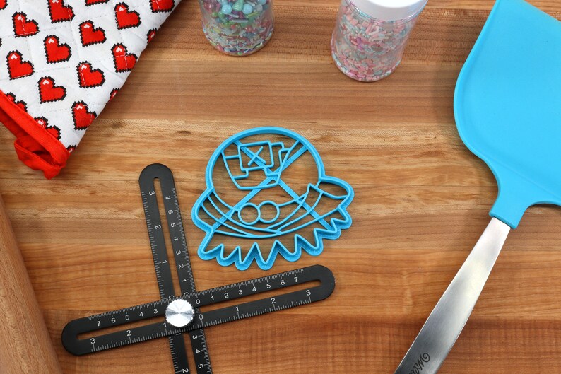 May include: A blue plastic cookie cutter in the shape of a character, with a spatula, jars of sprinkles, a red and white oven mitt, and a black measuring tool on a wooden surface. Baking and kitchen tools.