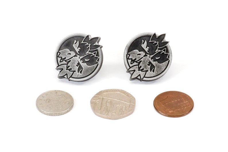 Op de afbeelding: Twee zilverkleurige metalen pinnen met een zwart ontwerp van een gestileerde bloem. De pinnen zijn rond en hebben een licht getextureerd oppervlak. De pinnen worden getoond naast drie munten.