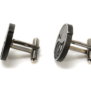 May include: A pair of black cufflinks with a circular design. The cufflinks have a silver metal backing.