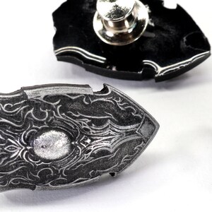 May include: Three silver-coloured metal pins shaped like shields with intricate designs. The pins have a black backing and a pin back.