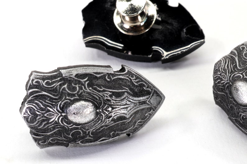 May include: Three silver-coloured metal pins shaped like shields with intricate designs. The pins have a black backing and a pin back.