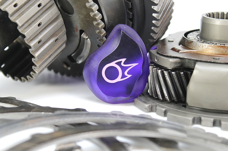 May include: A close-up of a purple glass gear shift knob with a white flame design. The knob is surrounded by metal gears.