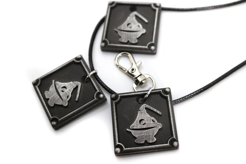 May include: Three silver-colored pendants with a black background and a cartoon character design. The pendants are attached to a black cord necklace with a silver clasp.