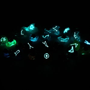 May include: A collection of glowing dice with constellations printed on each face. The dice are a variety of colors, including green, blue, and purple. The constellations are visible in the dark.