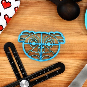 May include: Blue plastic cookie cutter shaped like an owl's face with large eyes and a beak. The cutter is on a wooden surface.