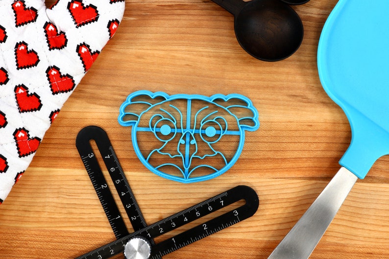 May include: Blue plastic cookie cutter shaped like an owl's face with large eyes and a beak. The cutter is on a wooden surface.