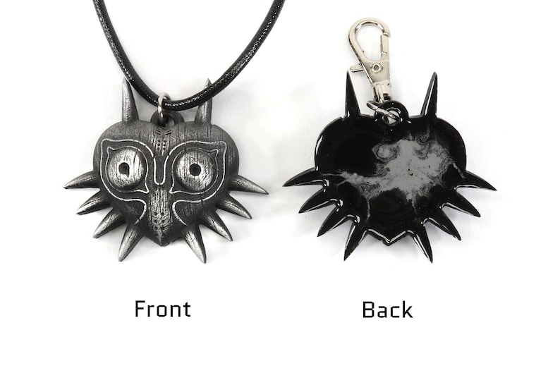 May include: A silver-colored Majora's Mask pendant necklace with a black cord. The back of the pendant is black. The pendant is also available as a keychain.