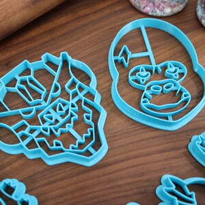 May include: Four blue plastic cookie cutters shaped like cartoon characters. The cutters are on a wooden surface.