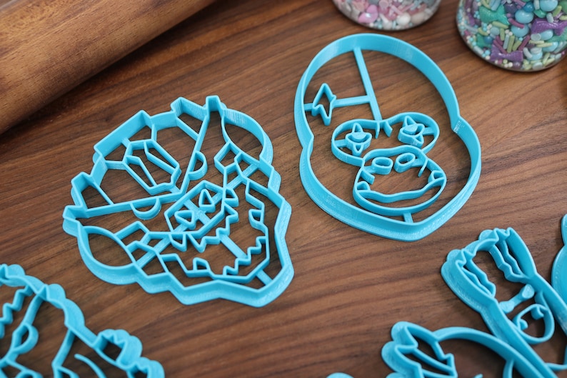 May include: Four blue plastic cookie cutters shaped like cartoon characters. The cutters are on a wooden surface.