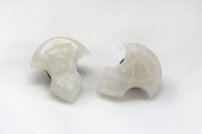 May include: Two white resin earrings with a crescent moon shape and a silver metal post. Each earring has a white design inside the crescent moon.