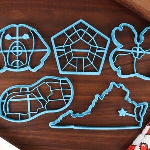 May include: A set of six blue plastic cookie cutters, including a dog, a house, a flower, a shoe, and the state of Virginia. The cutters are on a wooden surface.
